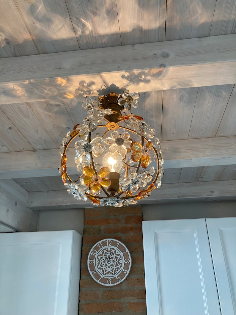 May include: A gold-toned chandelier with clear and amber glass flower accents hangs from a white wood ceiling. The chandelier is illuminated by a single light bulb.