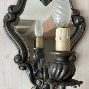 May include: Ornate, dark gray wall sconce with a mirror and a decorative, swirled light bulb. The sconce has a candle-like base and a curved arm. The mirror reflects the light bulb and the sconce's details.