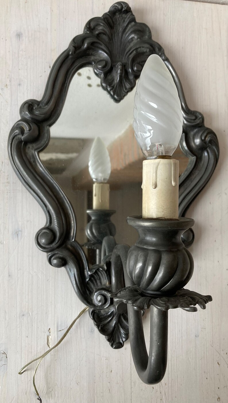 May include: Ornate, dark gray wall sconce with a mirror and a decorative, swirled light bulb. The sconce has a candle-like base and a curved arm. The mirror reflects the light bulb and the sconce's details.
