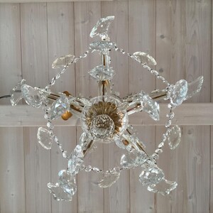 May include: A crystal chandelier with a gold-toned center and clear crystal drops. The chandelier is hanging from a white wooden ceiling.
