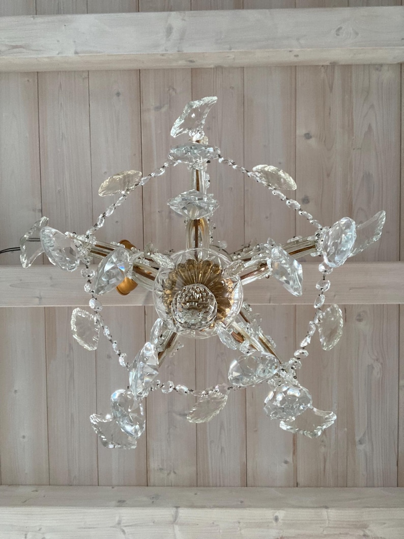 May include: A crystal chandelier with a gold-toned center and clear crystal drops. The chandelier is hanging from a white wooden ceiling.
