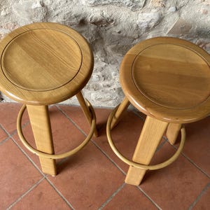 Op de afbeelding: Twee houten krukken met een lichtbruine afwerking. De krukken hebben een ronde zitting en een unieke, gebogen, vierpotige basis.