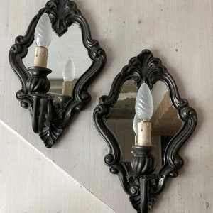 May include: Two ornate, dark gray wall sconces with small mirrors and decorative scrollwork. Each sconce holds a light bulb with a candle-like base. The sconces are mounted on a light-colored wall, creating a vintage aesthetic.
