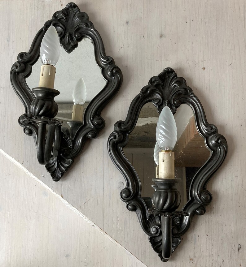 May include: Two ornate, dark gray wall sconces with small mirrors and decorative scrollwork. Each sconce holds a light bulb with a candle-like base. The sconces are mounted on a light-colored wall, creating a vintage aesthetic.