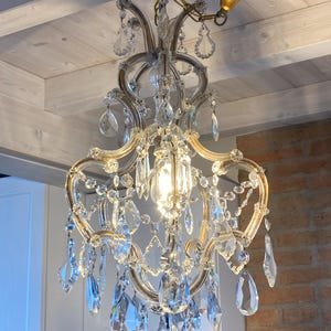 May include: A crystal chandelier with a gold finish and a chain hanging from the ceiling. The chandelier has multiple tiers of crystal drops and a central light bulb.