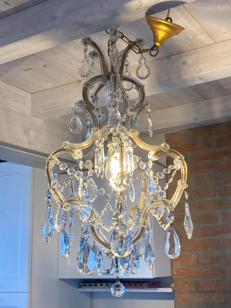 May include: A crystal chandelier with a gold finish and a chain hanging from the ceiling. The chandelier has multiple tiers of crystal drops and a central light bulb.