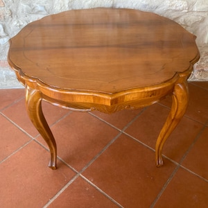 Pode incluir: Uma mesa oval de madeira vintage com borda ondulada. A mesa tem um acabamento castanho-médio quente e pernas curvas. A mesa está sobre um chão de ladrilhos de terracota.