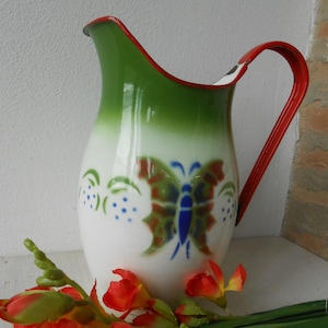 May include: A white enamel jug with a green top and red trim. The jug features a butterfly design in shades of brown, green, and blue. A red handle curves from the top to the side. Red and orange flowers are in the foreground.