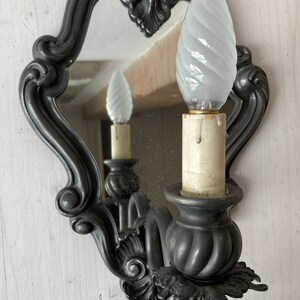 May include: An ornate, dark gray wall sconce with a small mirror and a white, swirled light bulb. The sconce has a decorative, scrolled design and a candle-like base. The mirror reflects the light bulb and the sconce's details.