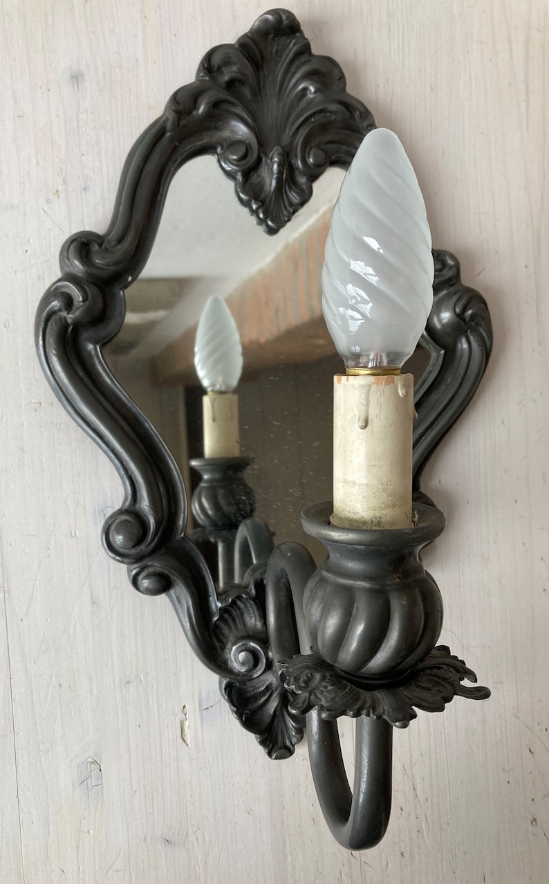 May include: An ornate, dark gray wall sconce with a small mirror and a white, swirled light bulb. The sconce has a decorative, scrolled design and a candle-like base. The mirror reflects the light bulb and the sconce's details.