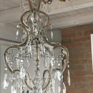 May include: A crystal chandelier with a gold finish and many crystal drops hanging from it. The chandelier is hanging from a ceiling with exposed wooden beams.