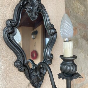 May include: An ornate, dark gray wall sconce with a decorative mirror and a single light bulb. The mirror has a scrolled, rococo-style frame. The light fixture features a decorative base and a swirled glass bulb.