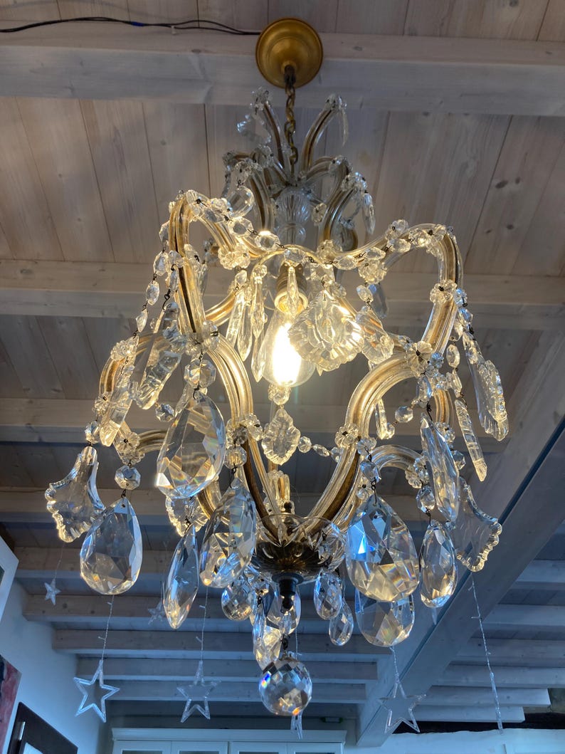 May include: A crystal chandelier with a gold frame and many hanging crystal drops. The chandelier is hanging from a wooden ceiling.