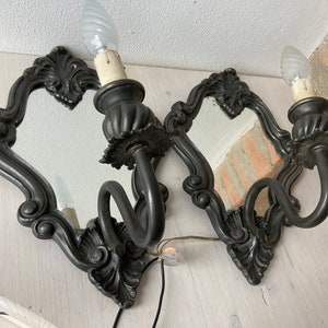 May include: Two ornate, dark gray wall sconces with small mirrors and candle-style light bulbs. The sconces have a decorative, scrolled design around the mirror and a curved arm holding the light fixture. The sconces are mounted on a light-colored wooden surface.