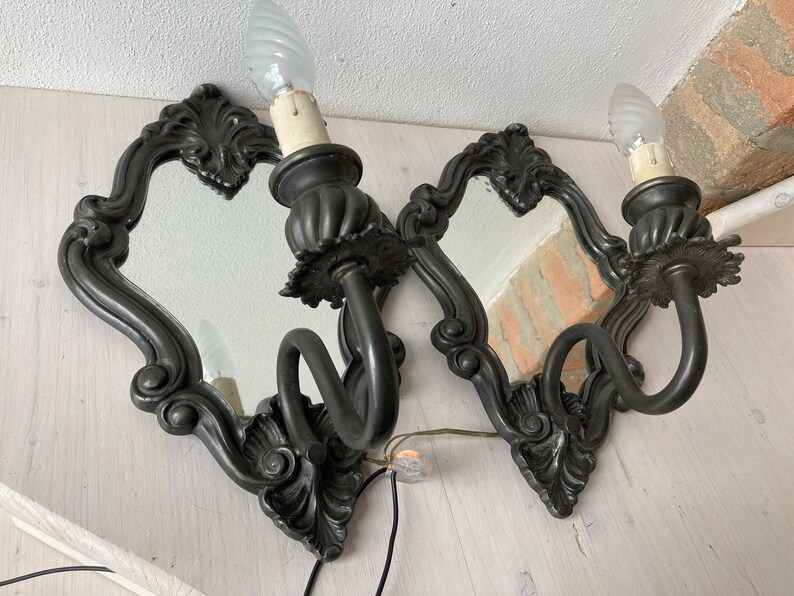 May include: Two ornate, dark gray wall sconces with small mirrors and candle-style light bulbs. The sconces have a decorative, scrolled design around the mirror and a curved arm holding the light fixture. The sconces are mounted on a light-colored wooden surface.