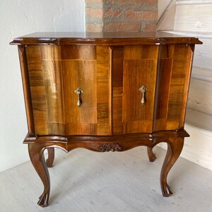 Puede incluir: Un gabinete de madera con acabado marrón pulido, que presenta dos puertas con paneles de veta vertical y tiradores de latón. La pieza se apoya sobre cuatro patas curvas tipo cabriolé con detalles tallados en el centro del faldón delantero.