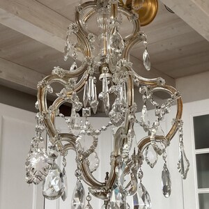 May include: A large, ornate crystal chandelier with a gold finish. The chandelier has multiple tiers of crystal drops and is hanging from a white ceiling.