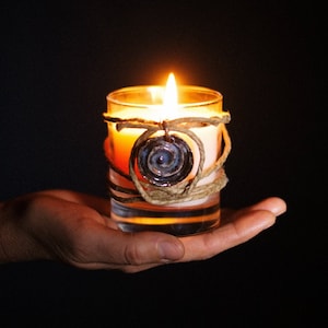 May include: A lit candle in a clear glass votive, held in an open hand against a black background. The candle is wrapped with twine and a decorative pendant. The flame glows brightly, illuminating the candle and its surroundings.
