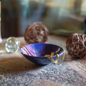 May include: A blue and purple ceramic bowl with a gold chain necklace inside. The bowl is sitting on a wooden table with a mosaic pattern.