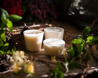 May include: Three ivory-colored votive candles with white wicks are arranged on a rustic wooden surface. Green leaves and sprigs of dried foliage surround the candles, creating a natural, earthy aesthetic. The candles are unlit, suggesting a focus on ambiance.