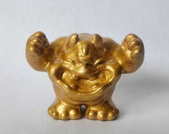 Op de afbeelding: Een klein, goudkleurig beeldje van een wezen met een brede, grijnzende mond, twee kleine hoorntjes op het hoofd en beide armen in de lucht met gebalde vuisten. Het figuurtje heeft een getextureerde, metallic afwerking en staat op twee benen.