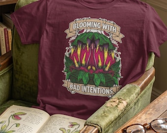 Op de afbeelding: Bordeauxrood t-shirt met een gedetailleerde afbeelding van een vleesetende plant met scherpe tanden en de tekst "BLOOMING WITH BAD INTENTIONS" op een banner. Het ontwerp bevat groene, roze en gele kleuren.