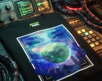 Op de afbeelding: Zwart t-shirt met een grafisch ontwerp met een groene, appelachtige planeet die in handen wordt gehouden tegen een blauwe en paarse achtergrond van de melkweg. De tekst "BEYOND PERIL" is boven de afbeelding gedrukt. Het shirt wordt gepresenteerd op een metalen oppervlak.