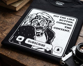 Op de afbeelding: Zwart t-shirt met een witte afbeelding van een schedelachtig figuur in een rechthoekige lijst. Het ontwerp bevat een duim-omhoog-pictogram, een bel en de woorden "SUBSCRIBE", "HIT THE LIKE BUTTON. CONSUME THE CONTENT."