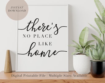 May include: A white framed print with black calligraphy that reads "there's no place like home". The print is on a wooden table with two potted plants.