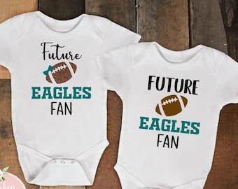 May include: Two white baby onesies with the text "Future Eagles Fan" in black and teal. Each onesie features a brown football graphic. The onesies are displayed on a wooden background.