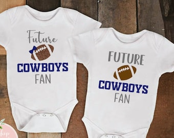 May include: Two white baby onesies with the text "Future Cowboys Fan" in gray and navy blue. A brown football graphic is above the text. The onesies are on a wooden background.