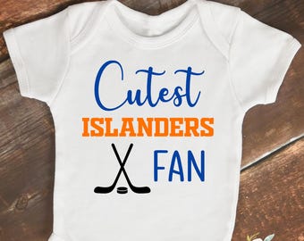 May include: White baby onesie with the text "Cutest Islanders Fan" in blue and orange. Two crossed hockey sticks are below the text. The onesie is on a wooden surface.