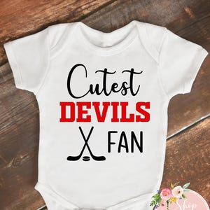 May include: White baby onesie with the text "Cutest DEVILS FAN" in black and red. Two crossed hockey sticks are below the text. The onesie is on a wooden background.