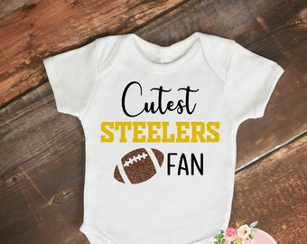 Könnte beinhalten: Weißer Baby-Body mit dem Text "Cutest Steelers Fan" in schwarzer und goldener Schrift und einer braunen Football-Grafik. Der Body wird auf einer Holzoberfläche präsentiert.