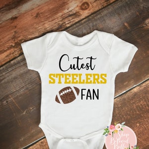 May include: White baby onesie with the text "Cutest Steelers Fan" in black and gold lettering, and a brown football graphic. The onesie is displayed on a wooden surface.