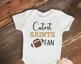 May include: White baby onesie with the text "Cutest SAINTS FAN" in black and gold lettering, and a brown football graphic. The onesie is on a wooden surface.
