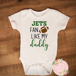 May include: White baby onesie with the text "JETS FAN LIKE MY daddy" in green and black. A brown football graphic is included. The onesie is displayed on a wooden surface.