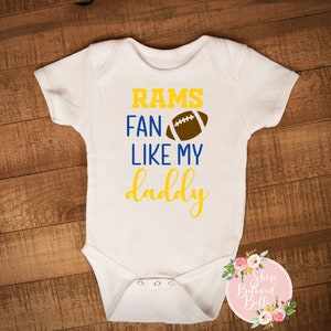 Puede incluir: Body de bebé blanco con el texto "RAMS FAN LIKE MY DADDY" en amarillo y azul, con un gráfico de balón de fútbol marrón. El body tiene cierre de corchetes y mangas cortas. Un logotipo floral está en la esquina inferior derecha.