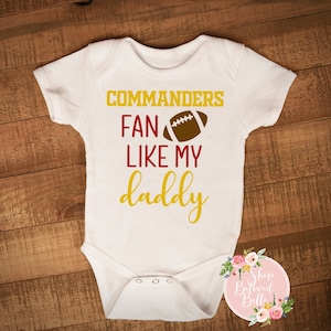 May include: White baby onesie with the text "Commanders Fan Like My Daddy" in gold and burgundy. A brown football graphic is above the word "Like". The onesie is on a wooden surface.