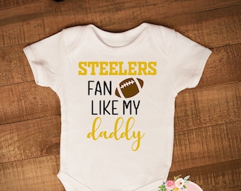 Könnte beinhalten: Weißer Baby-Body mit dem Text "Steelers Fan Like My Daddy" in Gold und Schwarz. Eine braune Football-Grafik befindet sich über den Worten "Like My". Der Body liegt auf einer Holzoberfläche.