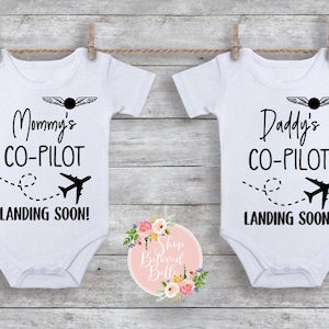 May include: Two white baby vests are suspended from a clothesline. One says "Mommy's CO-PILOT LANDING SOON!" and the other says "Daddy's CO-PILOT LANDING SOON!" Each features a pilot's wings and an aeroplane graphic.
