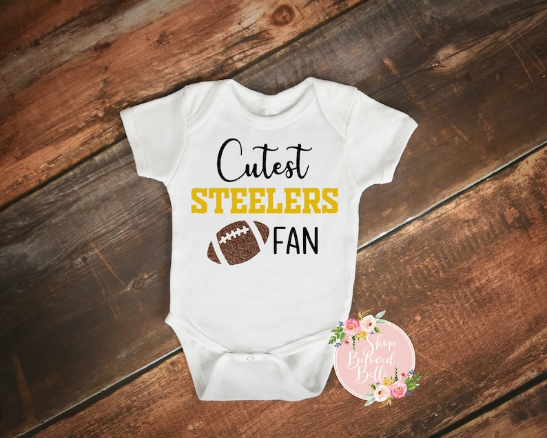 May include: White baby onesie with the text "Cutest Steelers Fan" in black and gold lettering, and a brown football graphic. The onesie is displayed on a wooden surface.