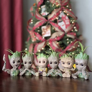 May include: Six small potted plants with green foliage and brown ceramic pots shaped like the character Groot from the Guardians of the Galaxy movie franchise. The pots are arranged in a row on a wooden surface.