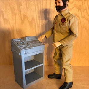 May include: A vintage action figure of a man in a tan uniform with a red and black emblem. The figure stands next to a gray control panel with a keyboard and buttons. The figure is wearing black shoes.