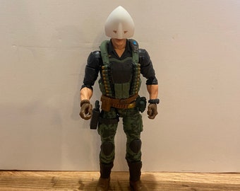 Puede incluir: Una figura de acción de camuflaje verde y negro que lleva una máscara blanca con la parte superior puntiaguda y dos agujeros para los ojos. La figura sostiene una pistola negra en su mano derecha y tiene un cinturón marrón con una hebilla plateada. La figura está de pie sobre una superficie de madera.