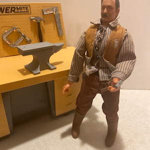 May include: A vintage toy figure of a man wearing a brown vest over a striped shirt, brown pants, and brown boots. He is holding a gun in his right hand. The figure is standing in front of a toy workbench with a small anvil and tools.
