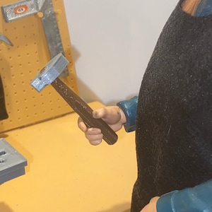 May include: A silver hammer with a brown wooden handle. The hammer is being held by a person wearing a blue shirt and a black apron.