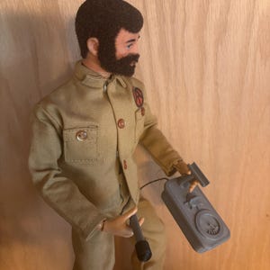 May include: A vintage action figure of a man in a tan uniform with a black beard and hair. The figure holds a gray device with a dial and a black microphone. The uniform has a red and black patch on the chest. The figure is wearing black boots.