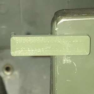 May include: A close-up of a light green plastic latch with a textured surface. The latch is attached to a larger object.