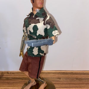 May include: A vintage action figure with a camouflage shirt, brown shorts, and a robotic arm holding a gun. The figure is standing on a wooden base.